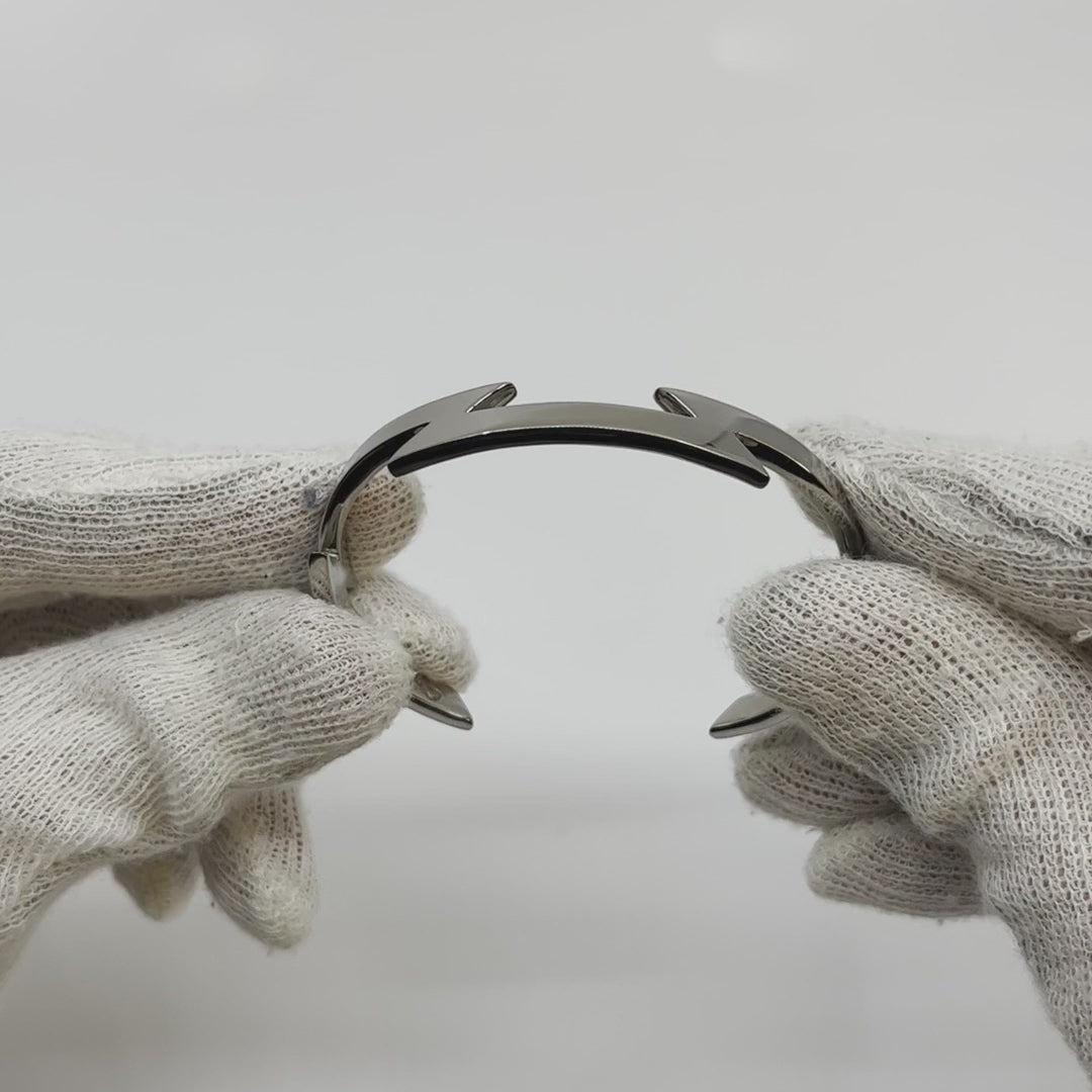 Video showing silver-colored titanium bangle with zig-zag pattern, 1 cm thick, being rotated to show all sides. Background is plain.