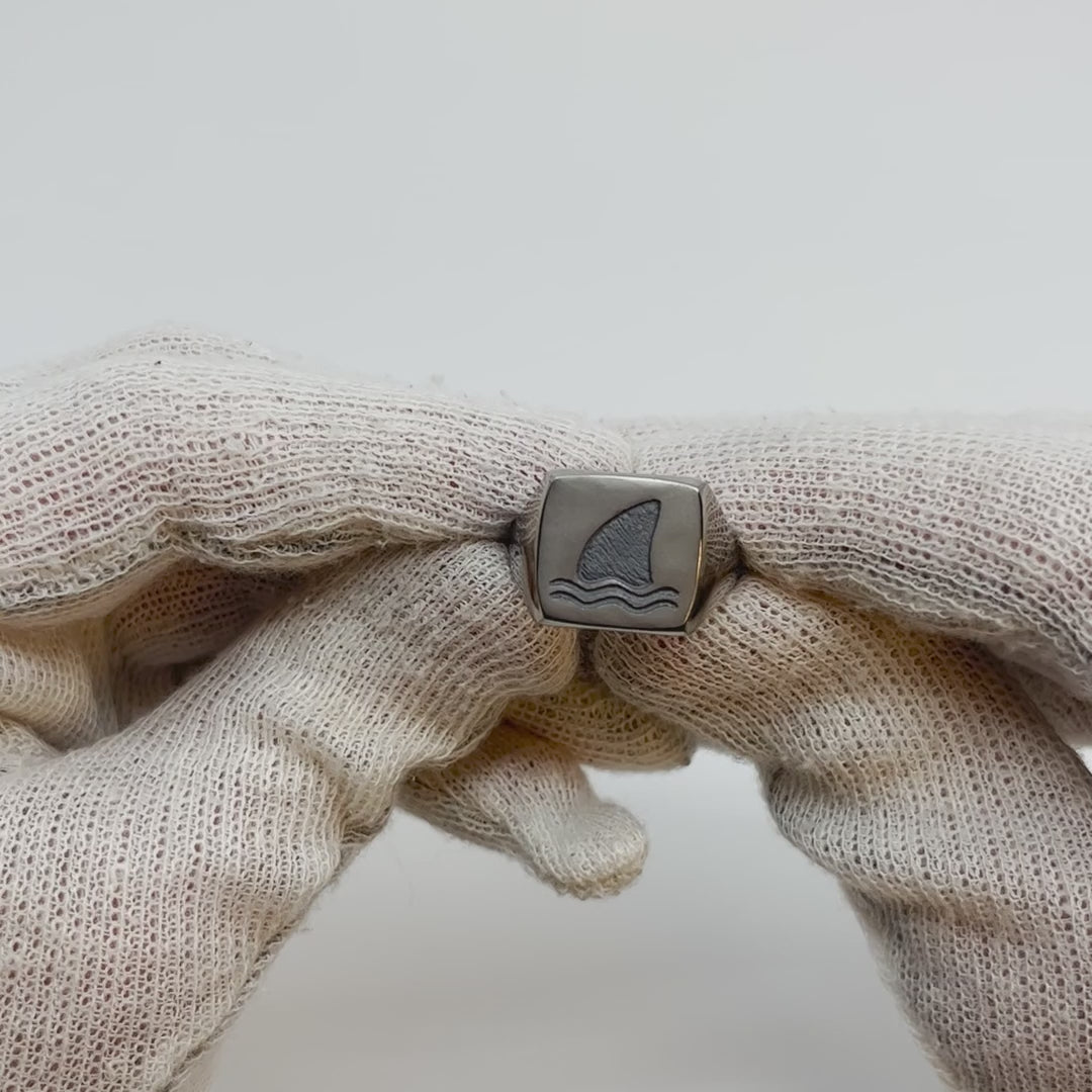 Showing single silver-colored titanium signet ring, 15x15 mm wide surface with an engraved shark fin from all angles. 